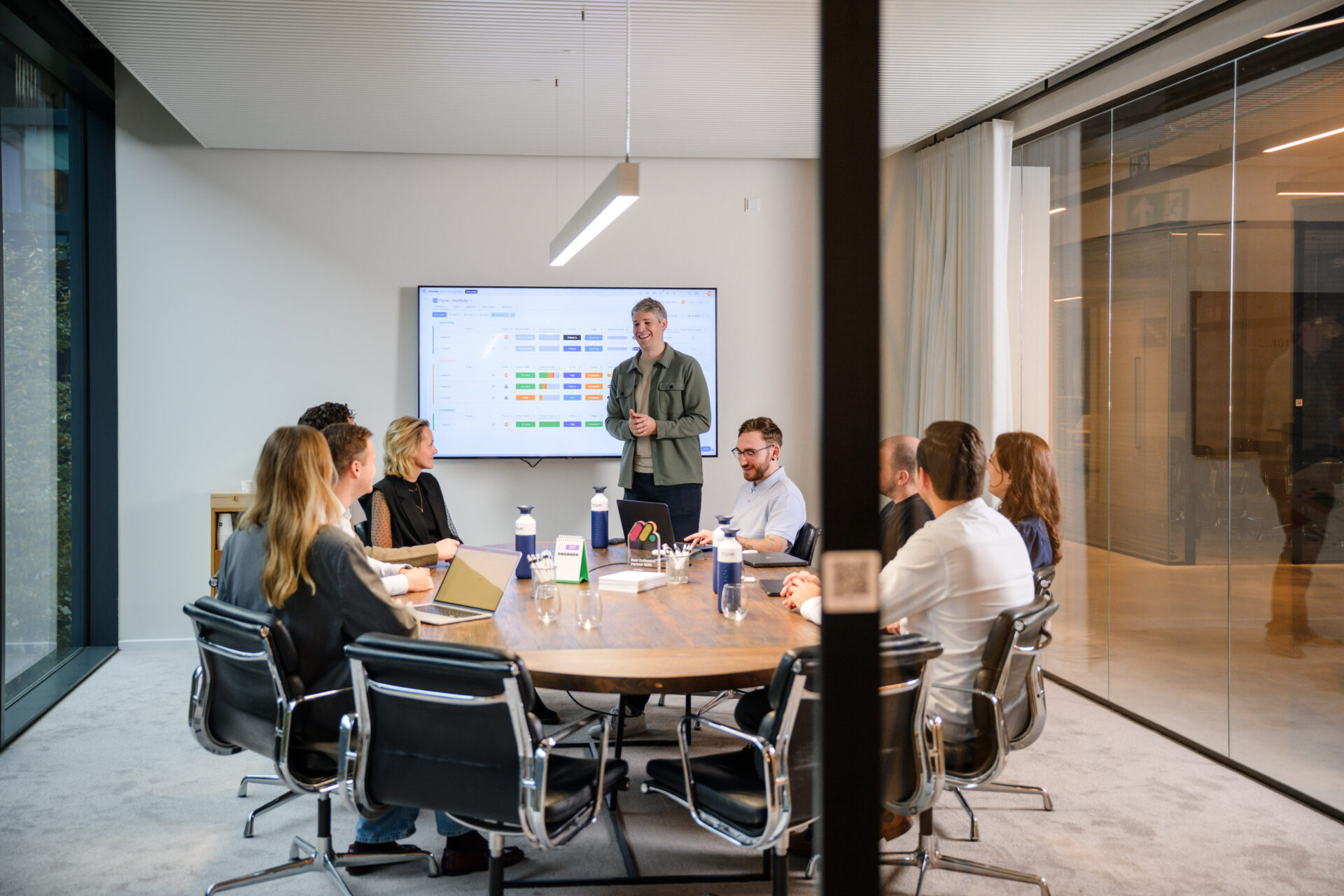 monday.com expert geeft een presentatie aan collega’s rond een ronde tafel in een moderne kantoorruimte.